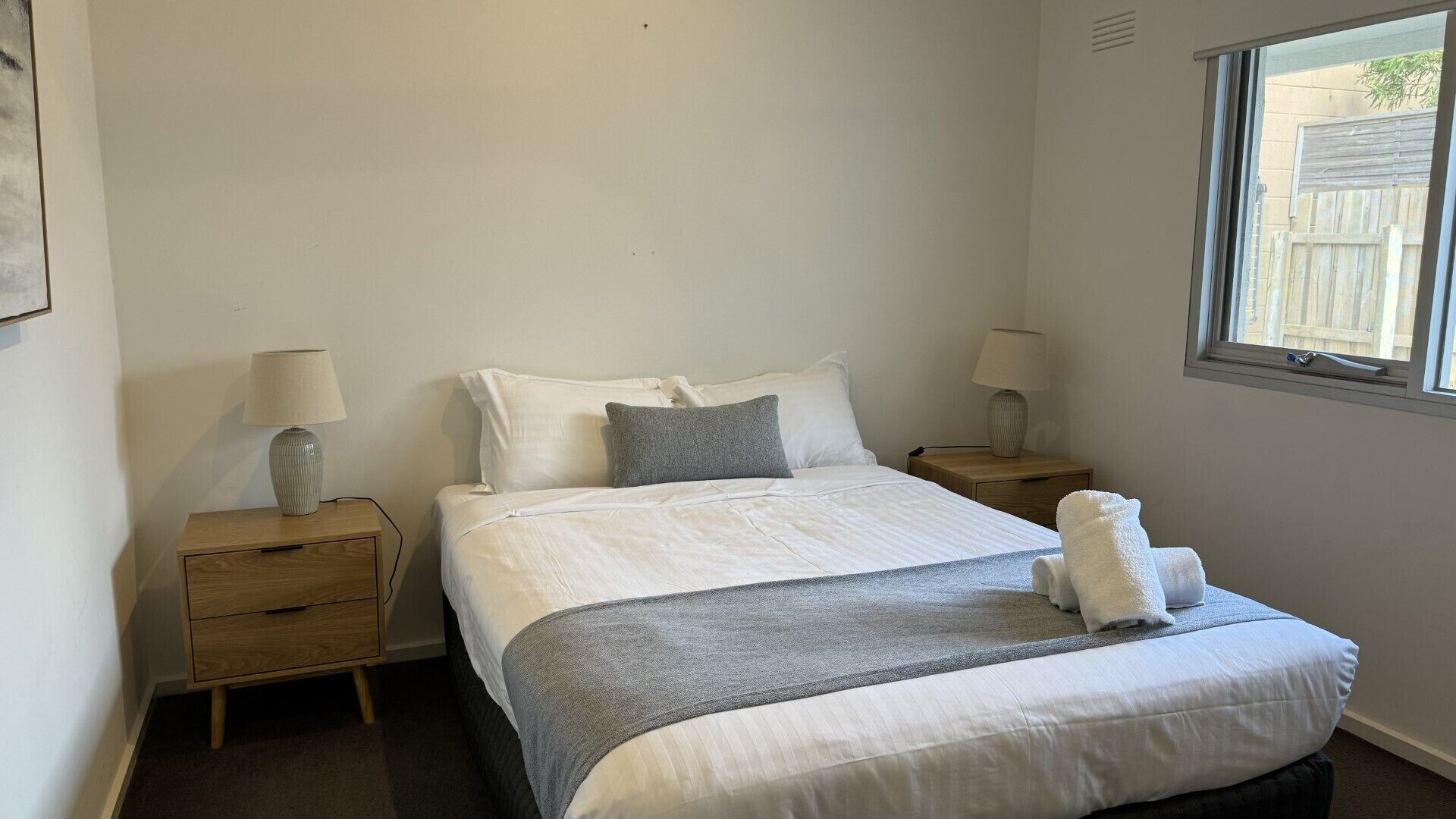 A neatly made bed with white and gray bedding, two bedside tables with lamps, and a window with a view of a fence.