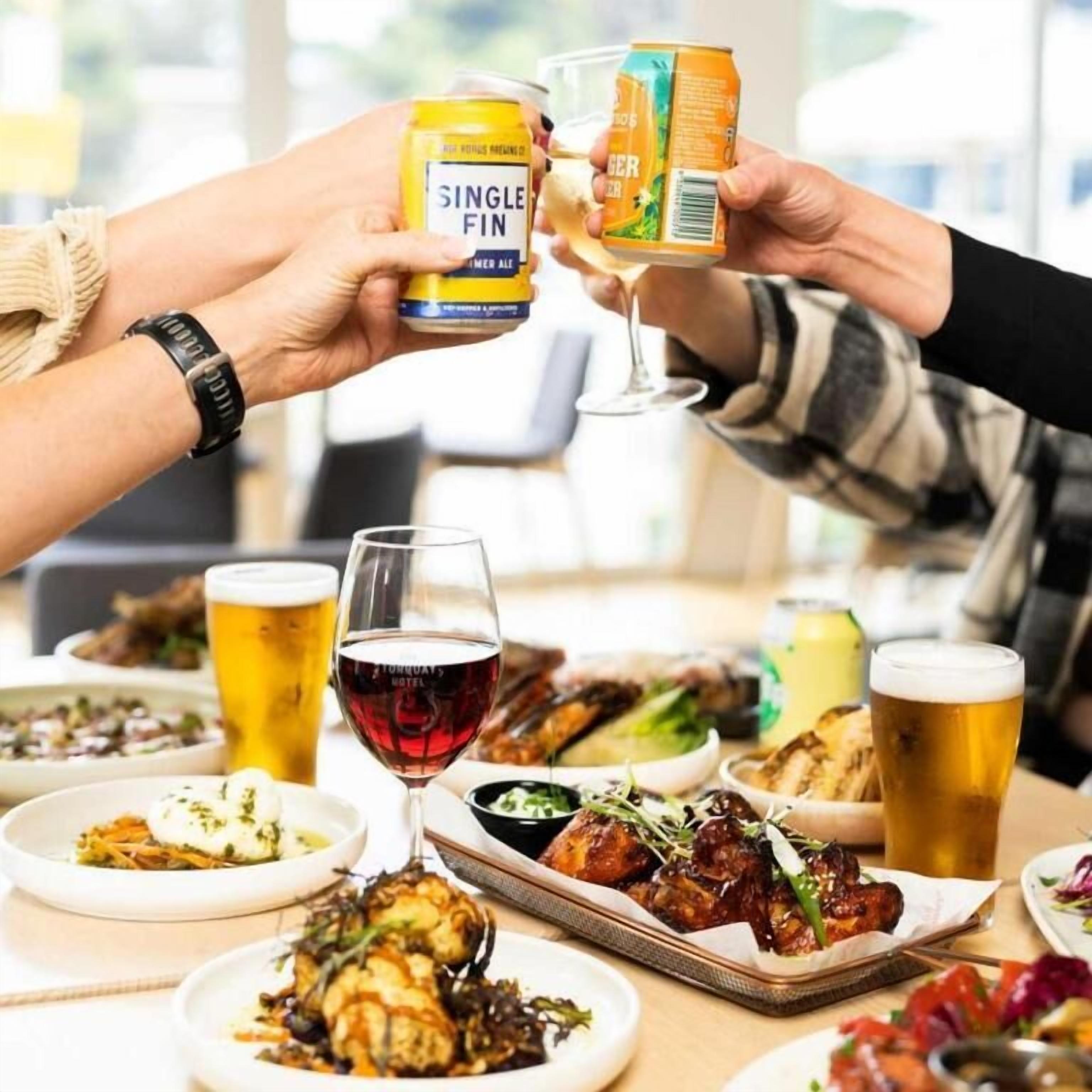 A group of people toasting with beer cans and glasses, surrounded by various dishes and drinks on a table.