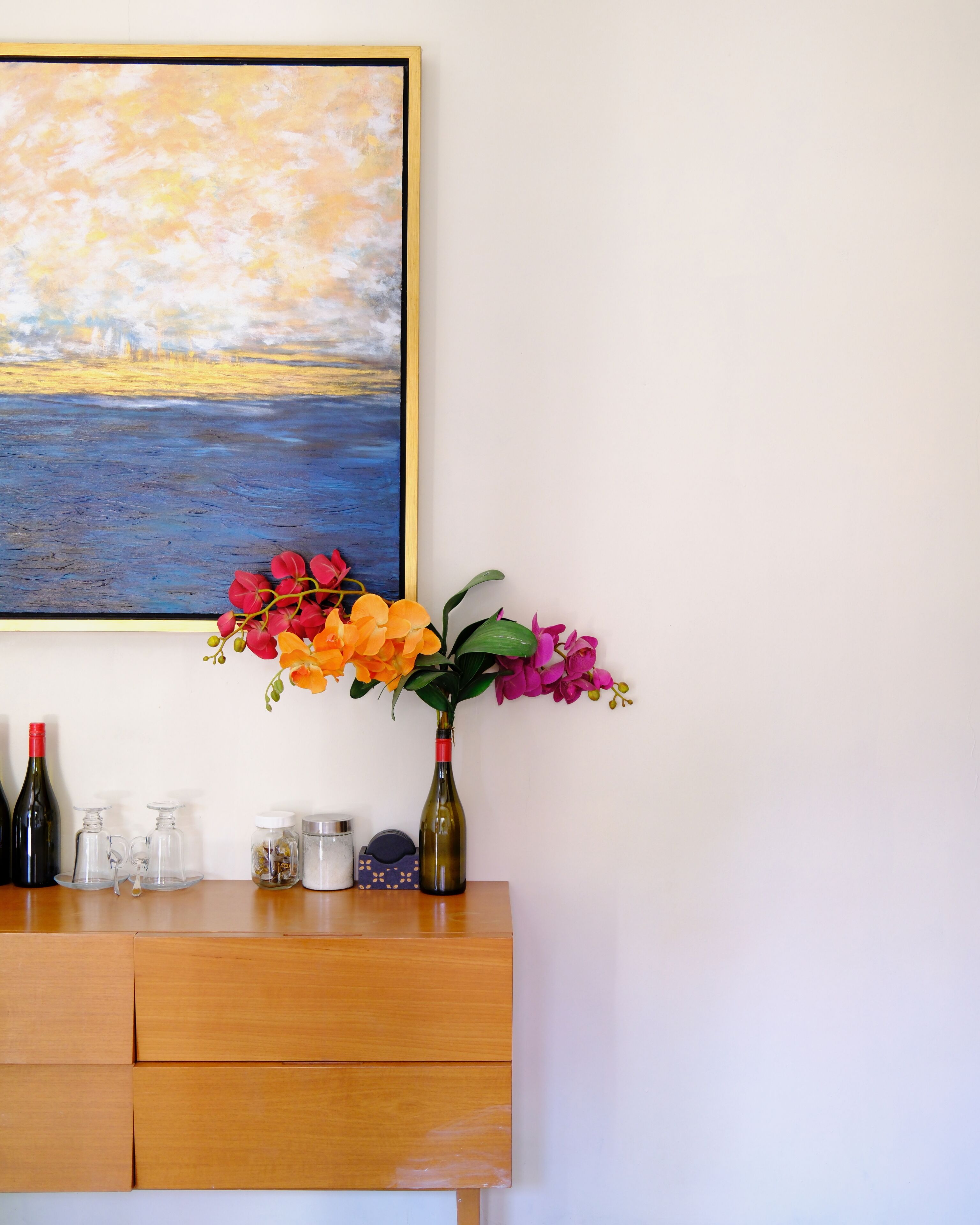 A wooden cabinet with a framed painting above it, a vase with flowers, and various glass containers.