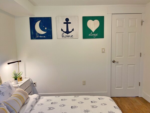 Un dormitorio con cama, una mesita de noche y una pared decorada con tres elementos: una luna y estrellas azules, un ancla y un corazón verde con la palabra 'always'.