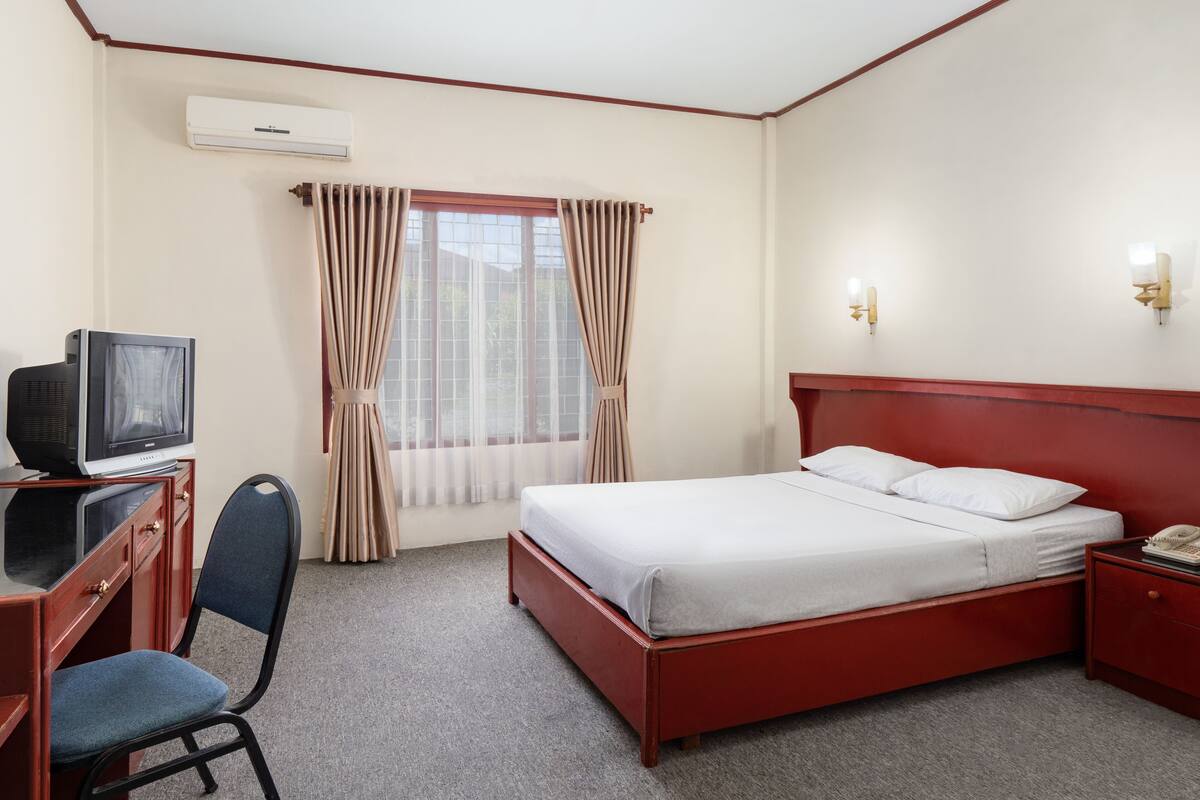Ein Hotelzimmer mit Bett, Schreibtisch mit Fernseher, Sessel, Nachttisch und einem fenster mit Vorhängen.