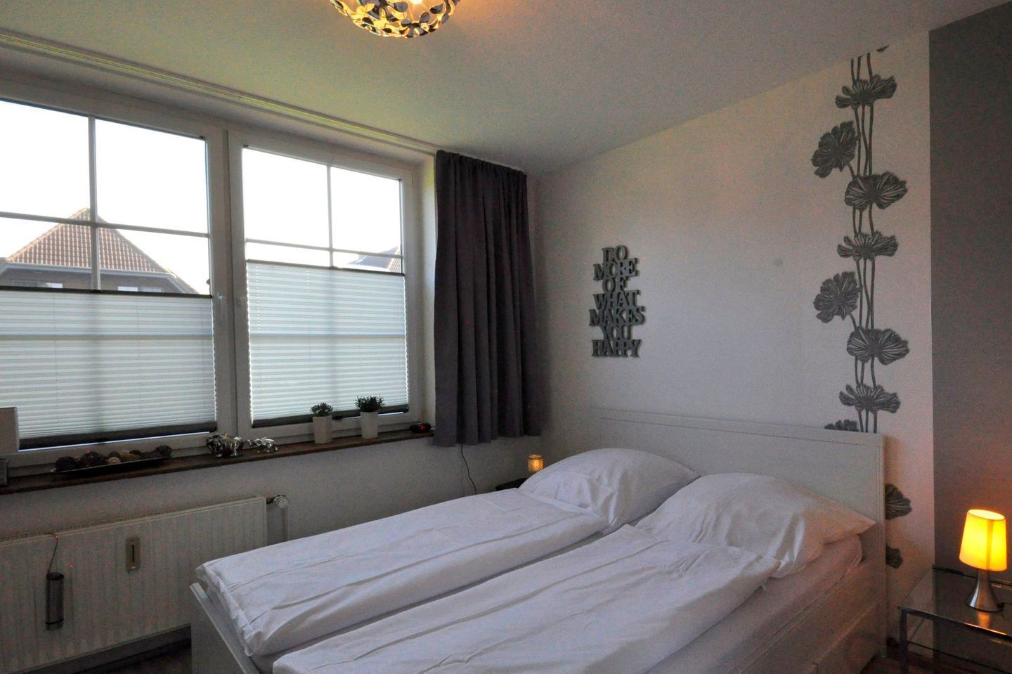 Une chambre avec deux lits, une fenêtre avec des stores, un radiateur, un élément de décoration murale et une lampe de chevet.