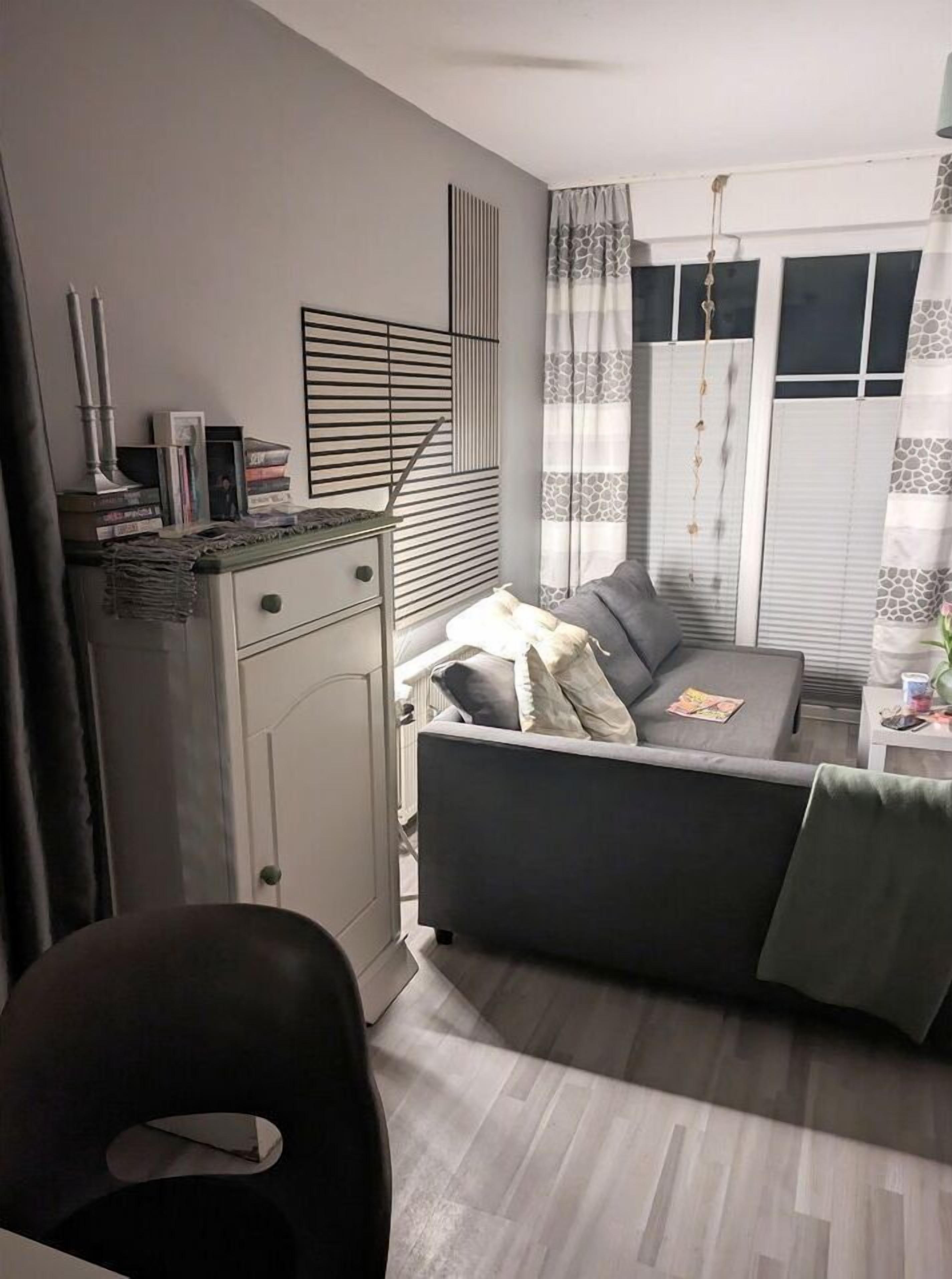 A modern living room with a grey sofa, a small table, and a cabinet. There is a window with curtains and a hanging plant.