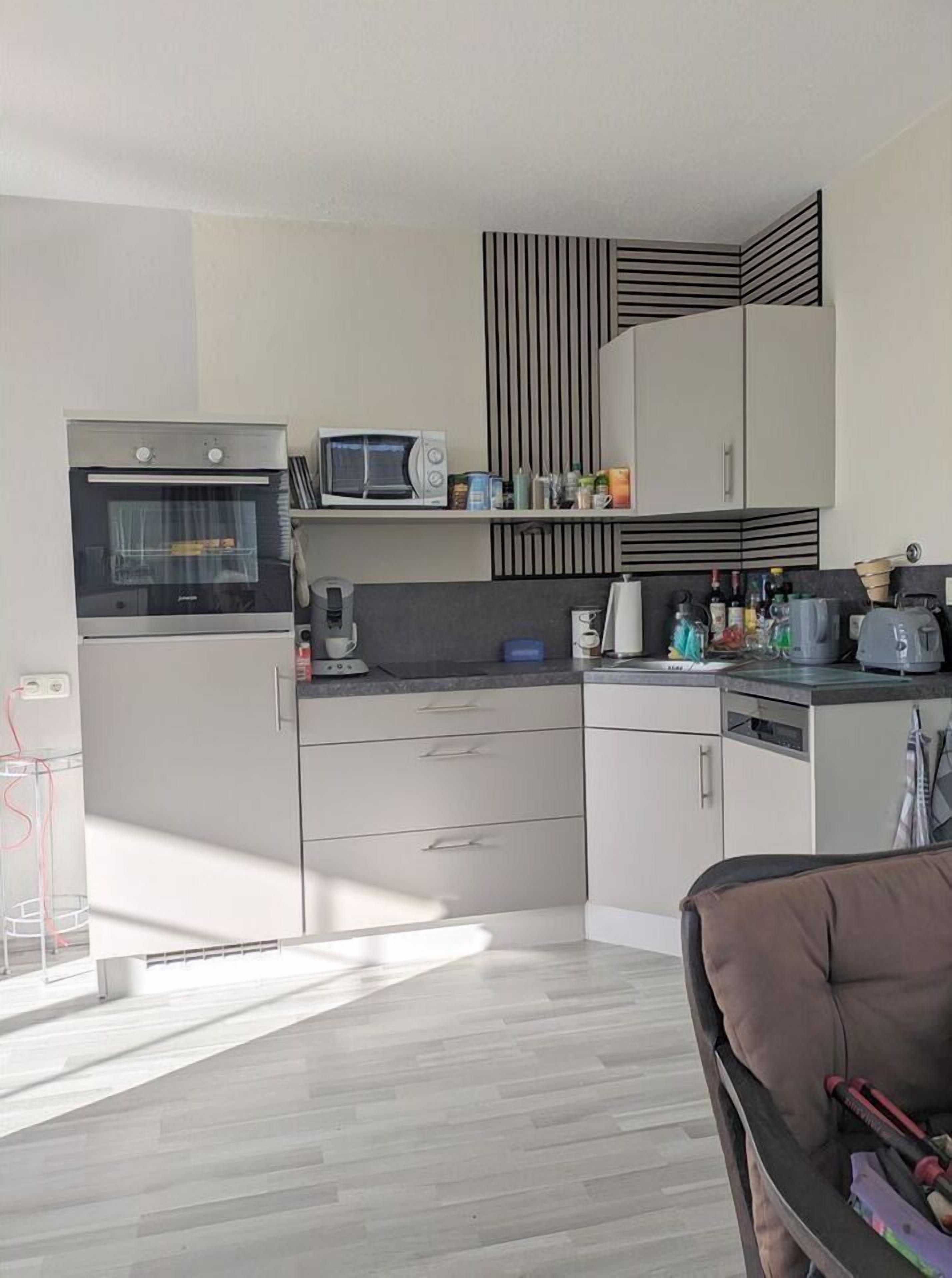 Een moderne keuken met een ingebouwde oven, magnetron en koffieapparaat. Er zijn kastjes, een aanrecht met diverse spullen, en een stoel op de voorgrond.