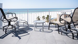 Ein Balkon mit drei Stühlen und einem kleinen Tisch mit Blick auf einen Strand bei blauem Himmel.