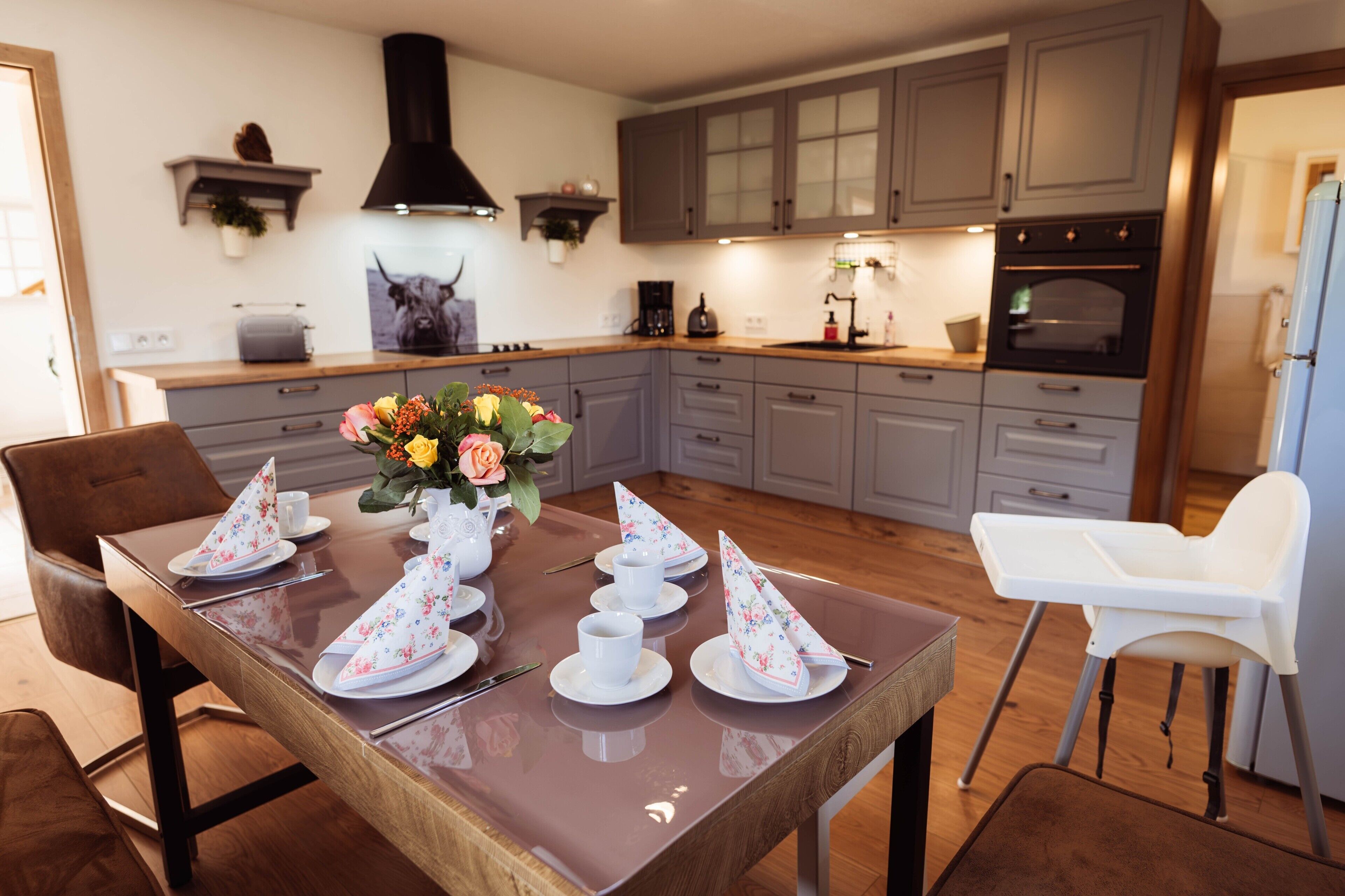 A modern kitchen with grey cabinets, a black range hood, and a dining area set for four with floral napkins and a vase of flowers.