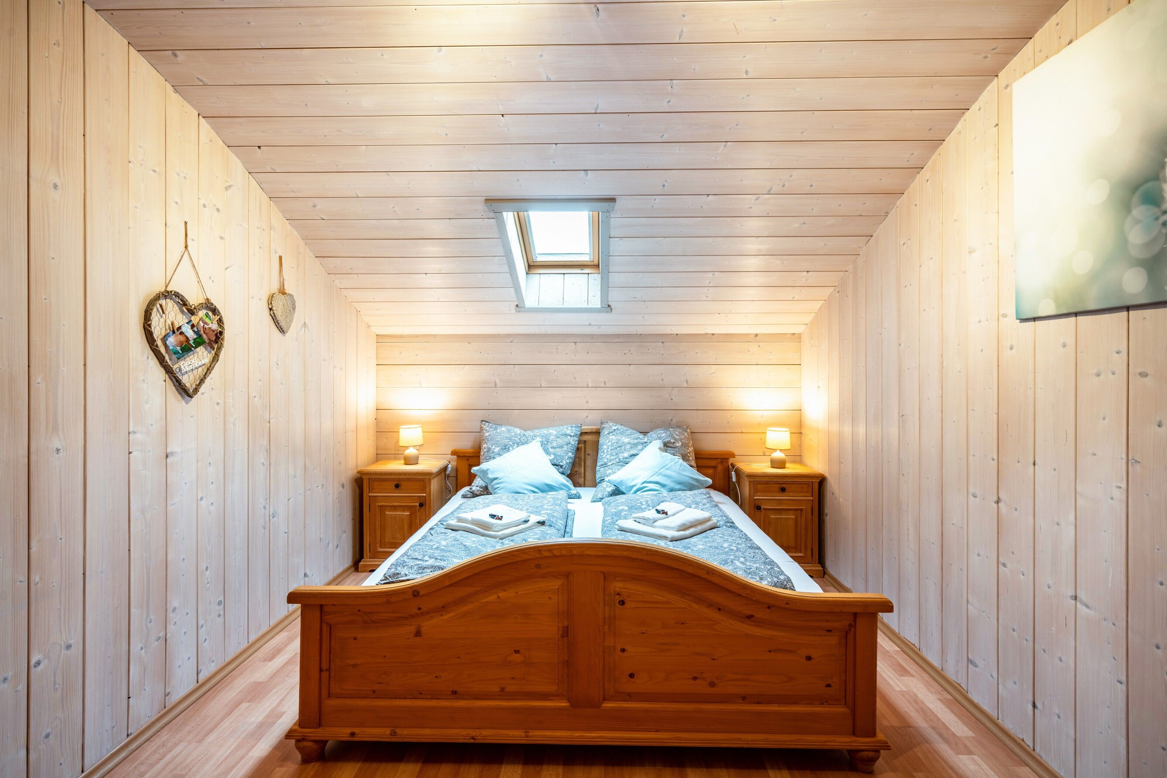Ein schmales Zimmer mit Holzbalkendecke, einem Oberlicht, einem Bett mit Holz-Kopfteil, zwei Nachttischen mit Lampen und einer herzförmigen Dekoration an der Wand.