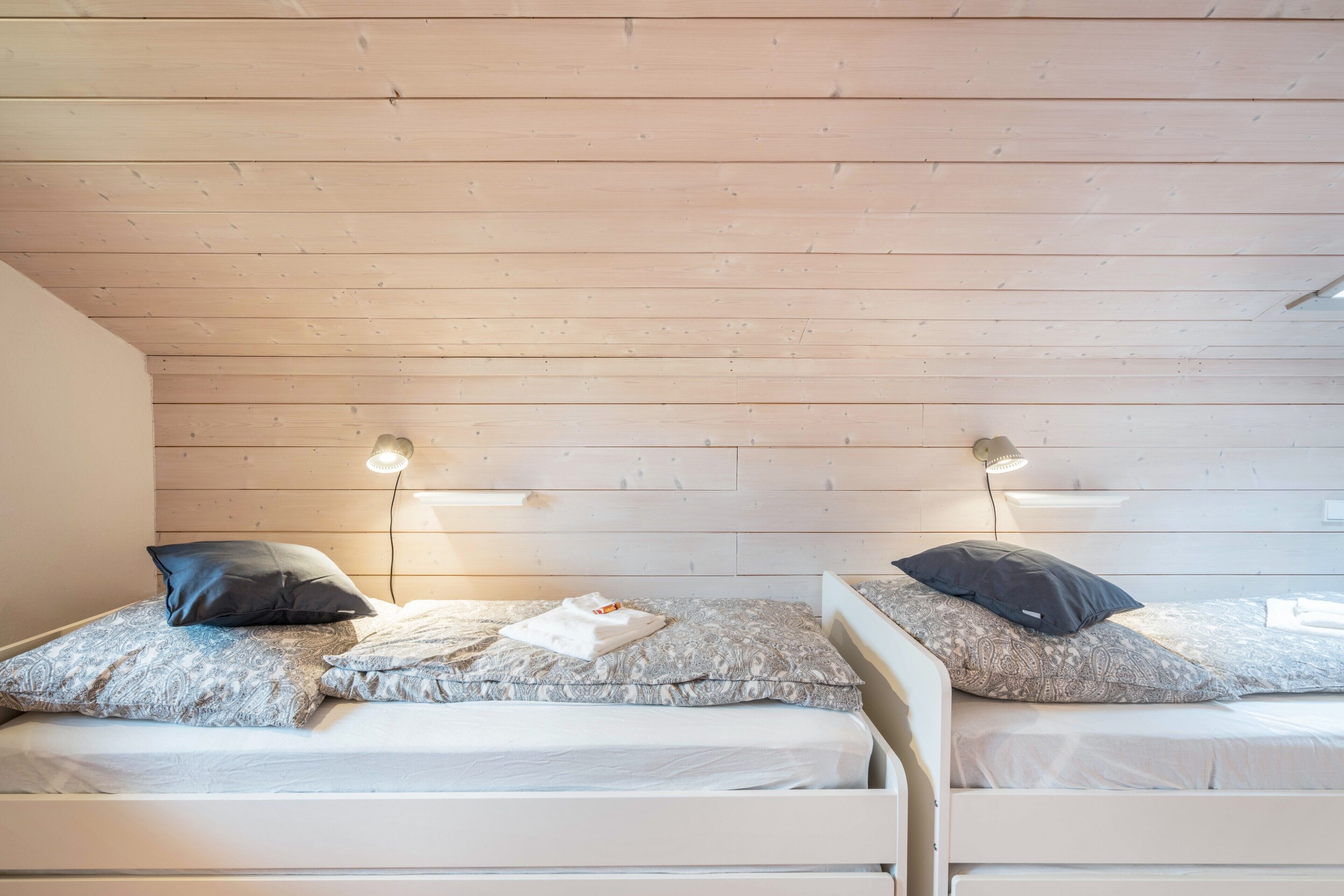 Une chambre avec deux lits, chacun agrémenté d’oreillers à motifs et recouvert d’une couverture blanche, sous un plafond en pente orné de panneaux de bois.