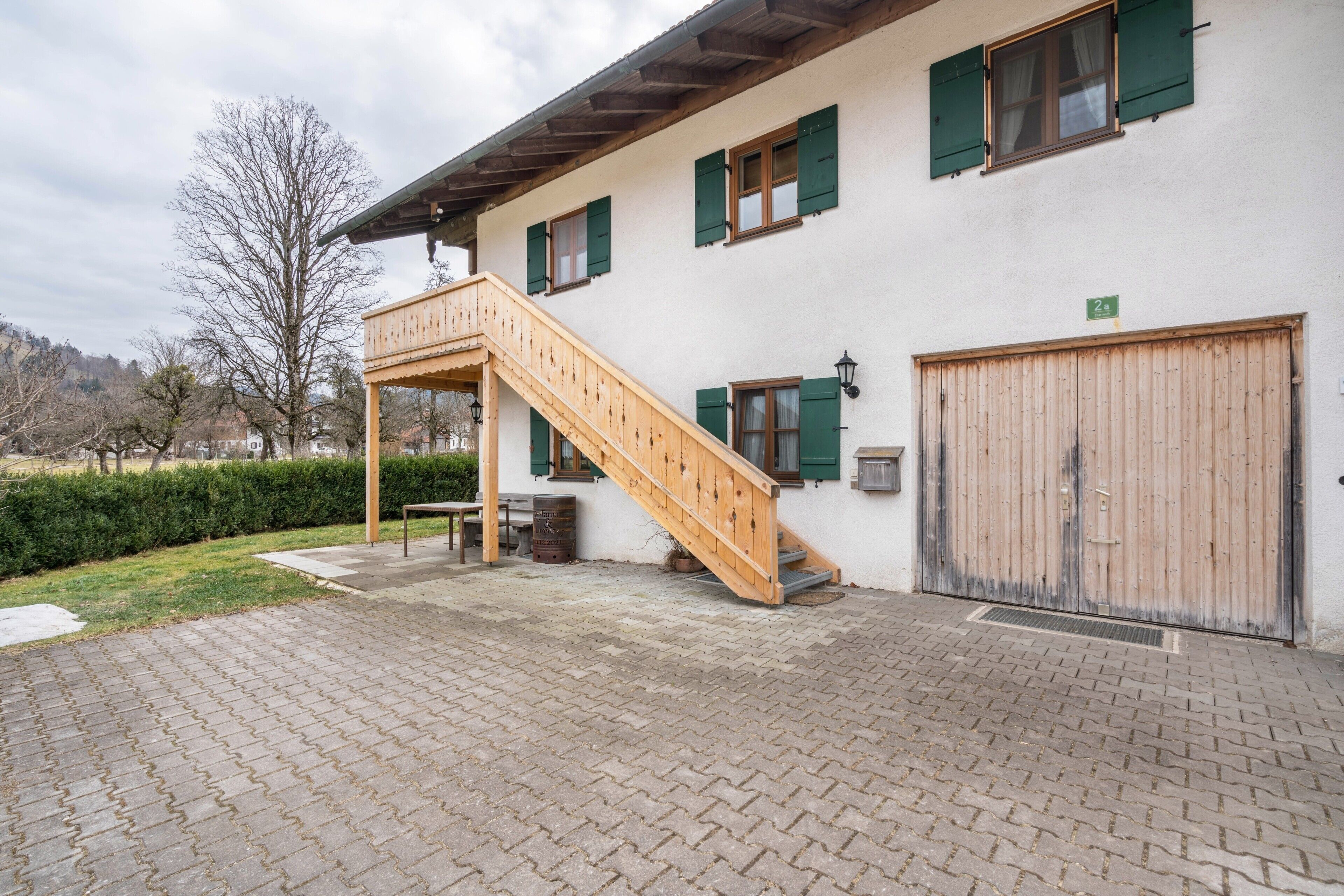 A building with a wooden staircase leading to a balcony, a large garage door, and a paved courtyard.