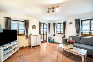 Una sala de estar con televisor, sofá, mesa de centro y ventanas con cortinas.