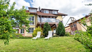 Ein zweistöckiges Haus mit einem Balkon, einer Rasenfläche mit zwei weißen Liegestühlen sowie verschiedenen Bäumen und Sträuchern.