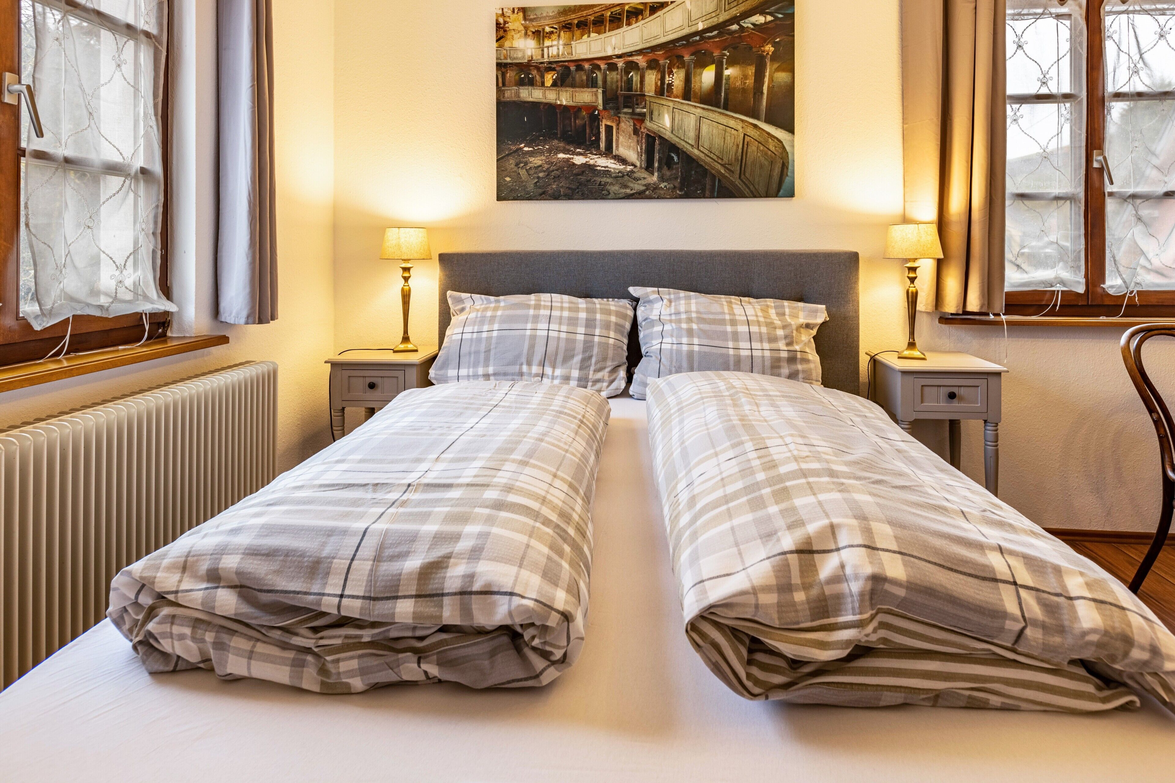 Een tweepersoonsbed met een geruit beddegoed, twee nachtkastjes met lampen, een radiator, en een schilderij van een oud theater aan de muur.