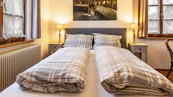 Een tweepersoonsbed met een geruit beddegoed, twee nachtkastjes met lampen, een radiator, en een schilderij van een oud theater aan de muur.
