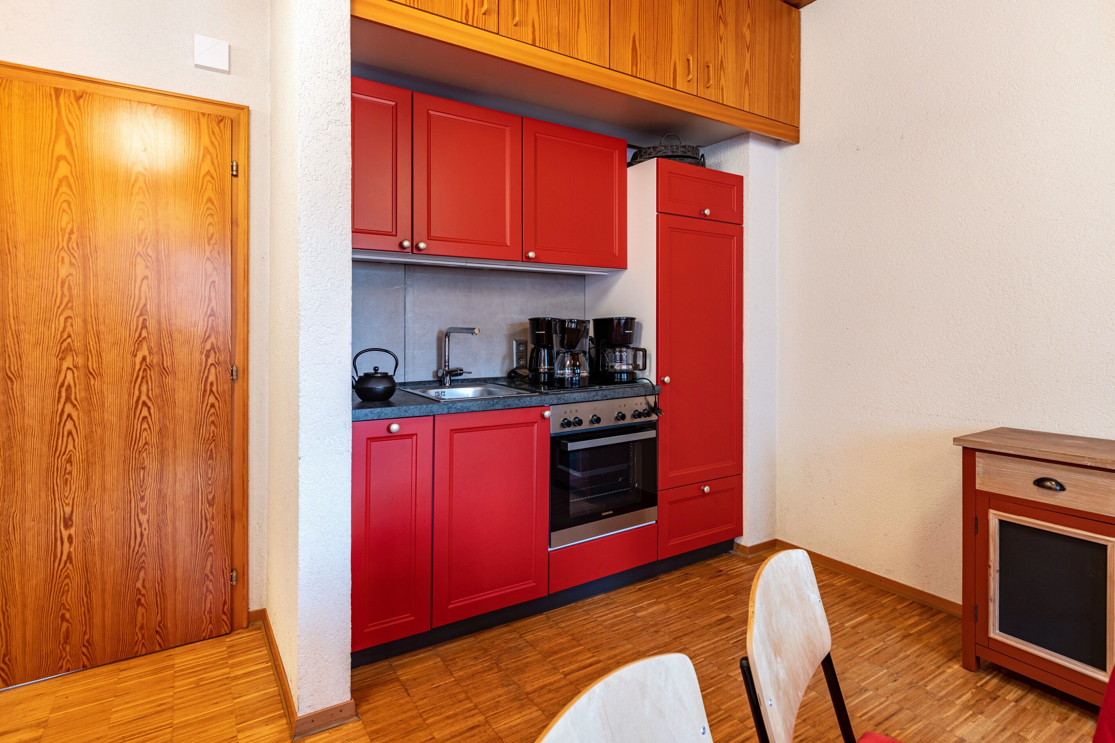 A compact kitchen with red cabinets, a built-in oven, and a small wooden cabinet.