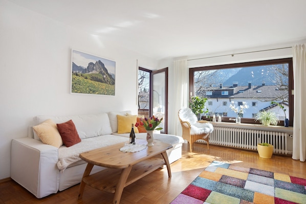 A modern living room with a white sofa, a wooden coffee table, a colorful rug, and a view of mountains through a large window.