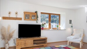 Un soggiorno con televisore a schermo piatto, mobile TV in legno, un tappeto colorato, una finestra vista alberi e una poltrona bianca.