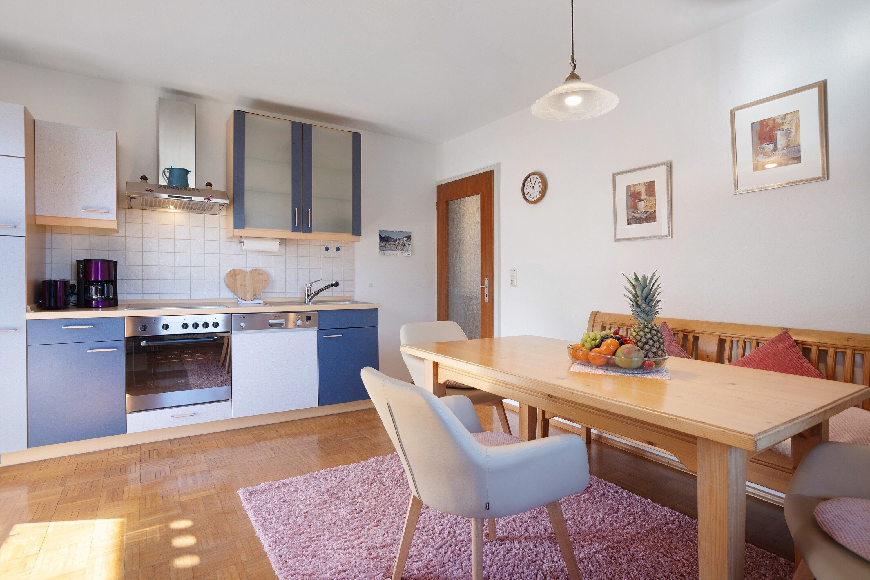 A modern kitchen with a dining area, wooden furniture, and a fruit bowl on the table.