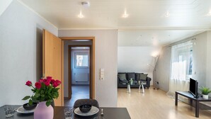 A modern living room with a sofa, a coffee table, and a TV. There's a dining area with a table set for two, a vase with flowers, and a hallway leading to another room. - Apartment 'Am Hirnach' with Shared Terrace, Shared Garden and Wi-Fi (Sindelfingen)