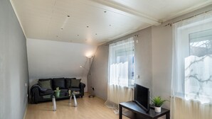 A living room with a sofa, a TV on a stand, a lamp, and a window with sheer curtains. - Apartment 'Am Hirnach' with Shared Terrace, Shared Garden and Wi-Fi (Sindelfingen)