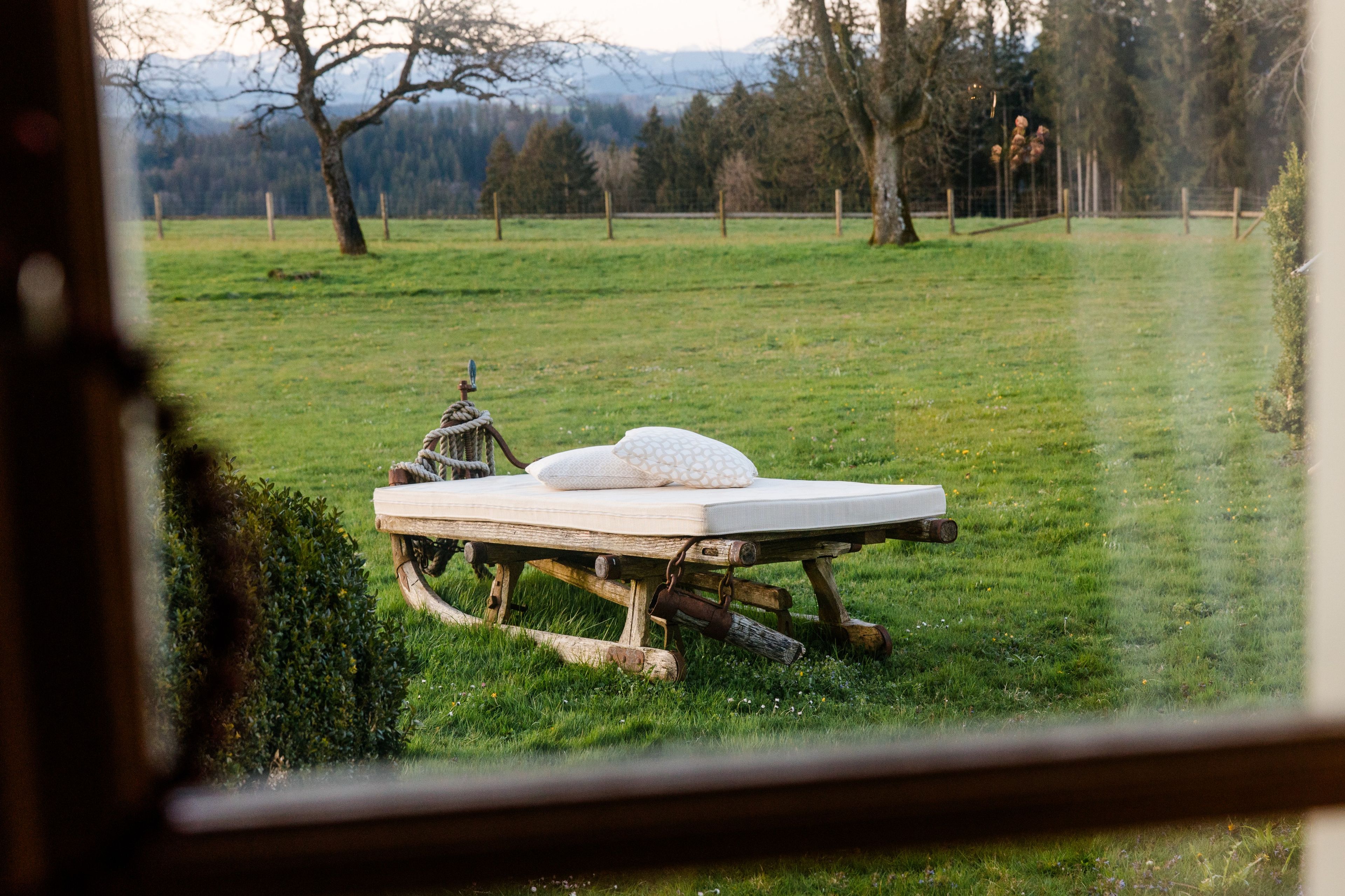 Un lit en bois rustique, avec un matelas et des oreillers, se trouve dans un champ d’herbe, vu à travers une fenêtre dont le cadre est en métal.