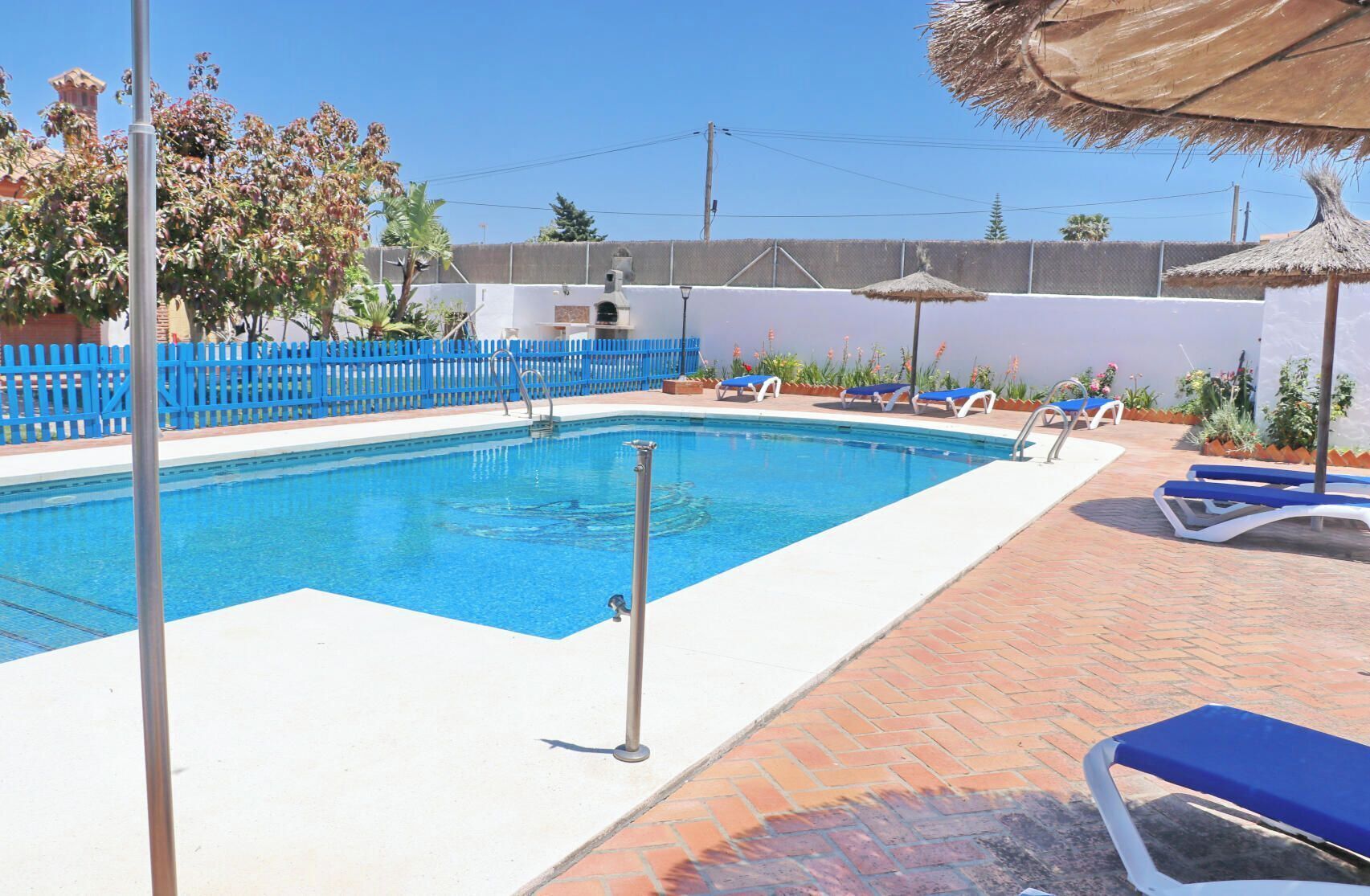 Piscina con agua azul, rodeada por un muro blanco y una cerca azul. Hay sillones de descanso y sombrillas alrededor de la piscina.