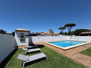 Piscina com espreguiçadeiras e um pérgola, cercada por um muro branco e uma árvore.