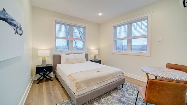A bedroom with a bed, two bedside tables, a chair, and a window with blinds. - Stay on Clay - Suite #6 (Muskegon)