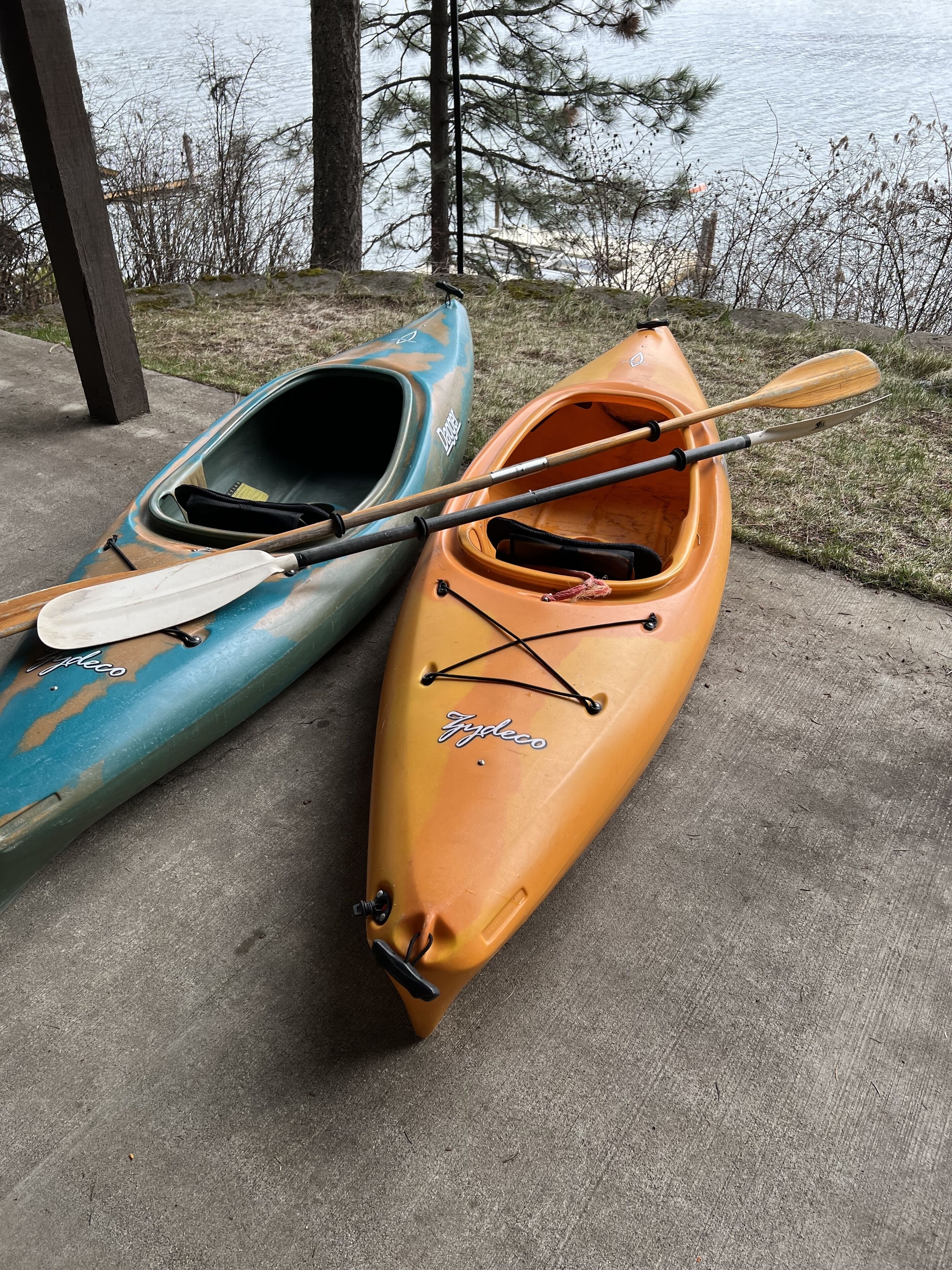 Dos kayaks, uno azul y uno naranja, con remos y cuerdas, sobre una superficie de hormigón cerca del agua.