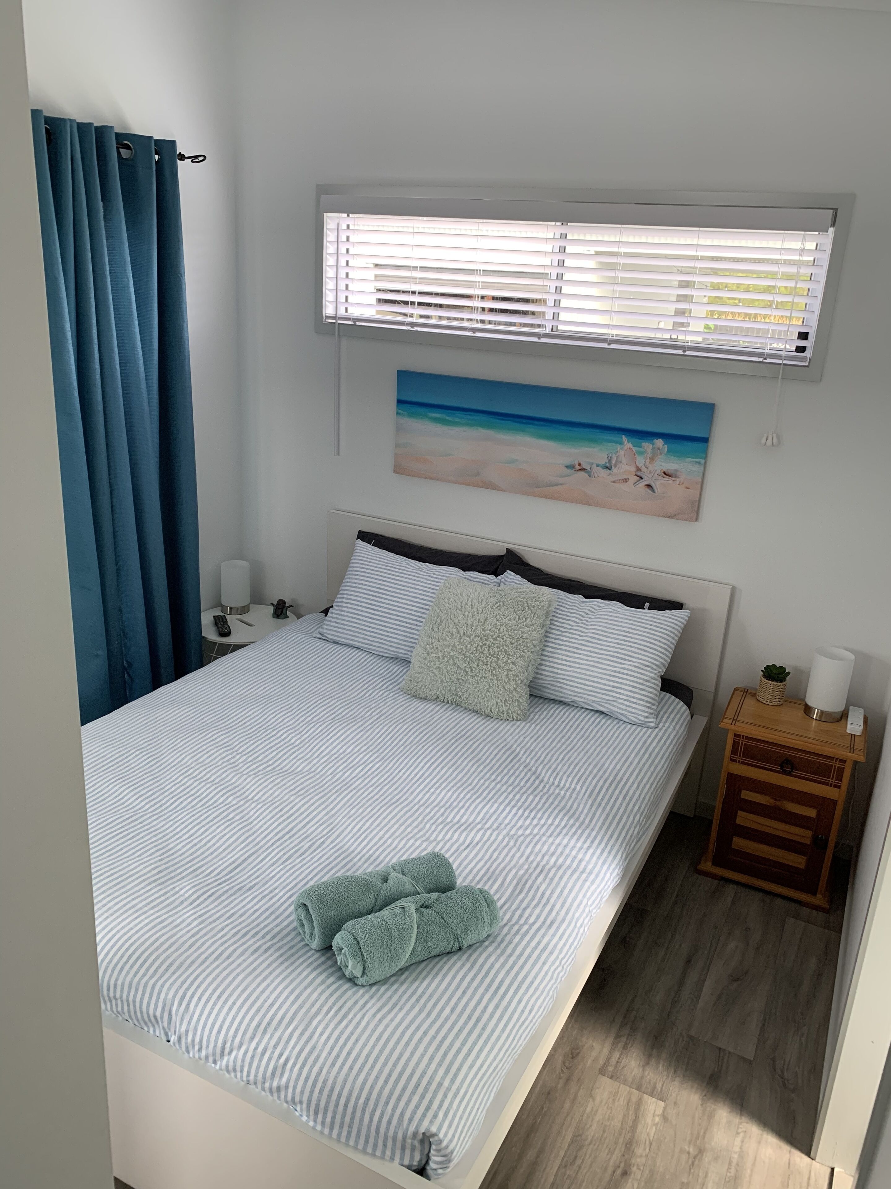 A neatly made bed with striped bedding, a painting of a beach scene, a bedside table with a lamp, and a window with blinds.