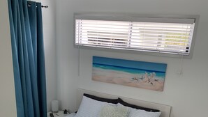 A neatly made bed with striped bedding, a painting of a beach scene, a bedside table with a lamp, and a window with blinds. - Peaceful Oasis at Cooloola Cove (Cooloola Cove)