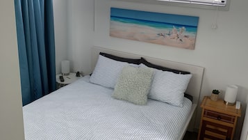 A neatly made bed with striped bedding, a painting of a beach scene, a bedside table with a lamp, and a window with blinds.
