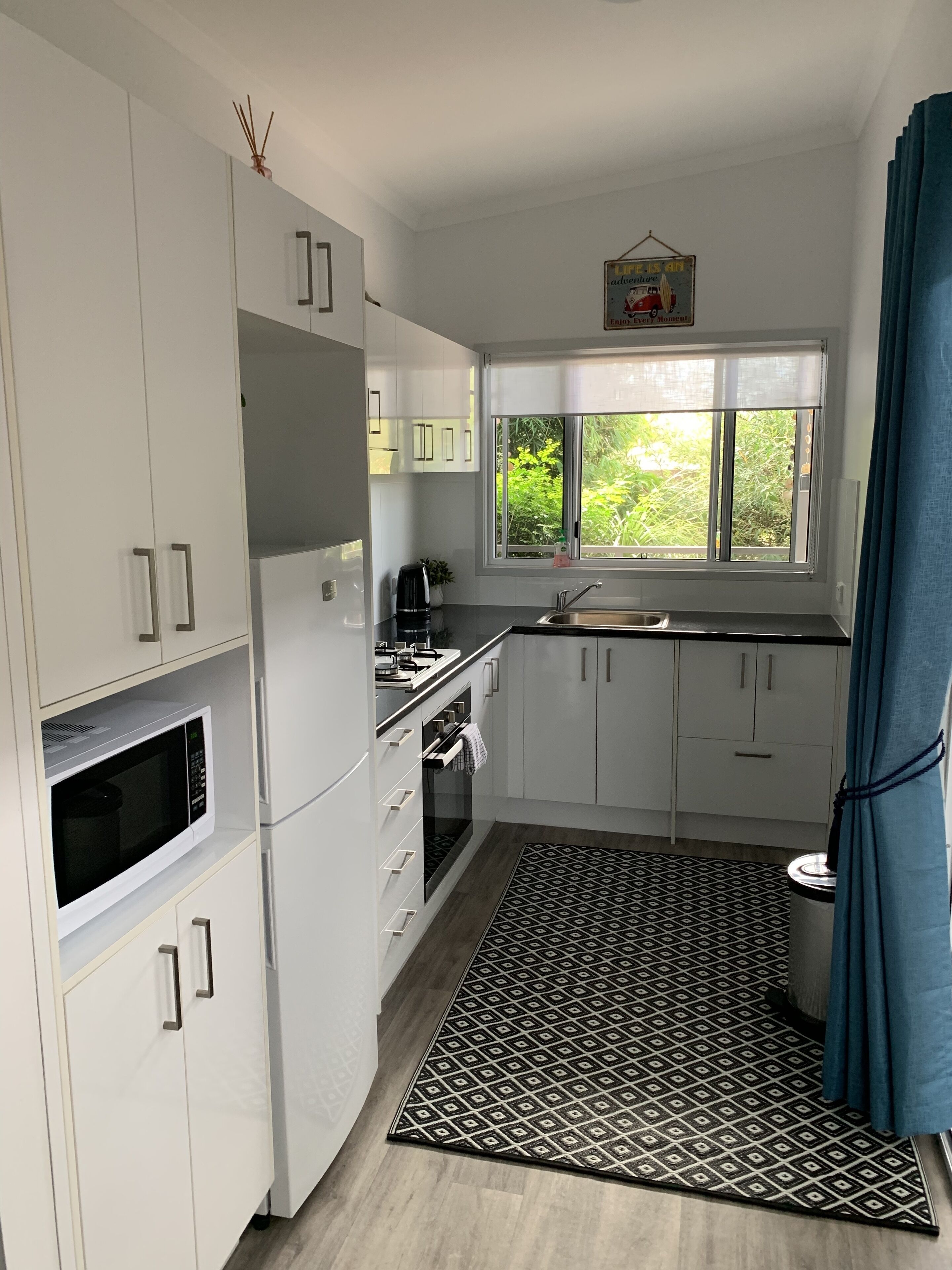 A compact kitchen with white cabinets, a microwave, a kettle, and a window with a view of greenery.