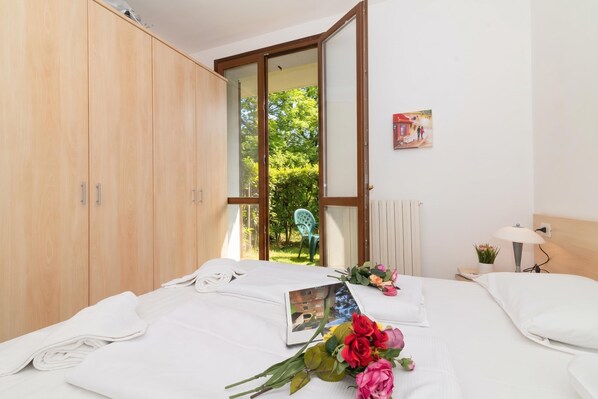 A hotel room with a bed, a wooden wardrobe, a window with a view of greenery, and a small table with a lamp.