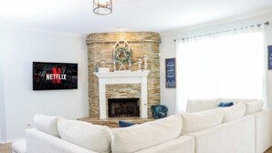 Un salon avec un canapé d’angle blanc, une cheminée en pierre, un logo Netflix et un mur orné d’une citation inspirante.