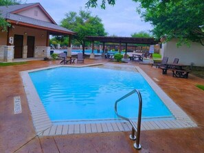 A swimming pool with a metal handrail, surrounded by lounge chairs and tables, with a covered area and a building in the background.