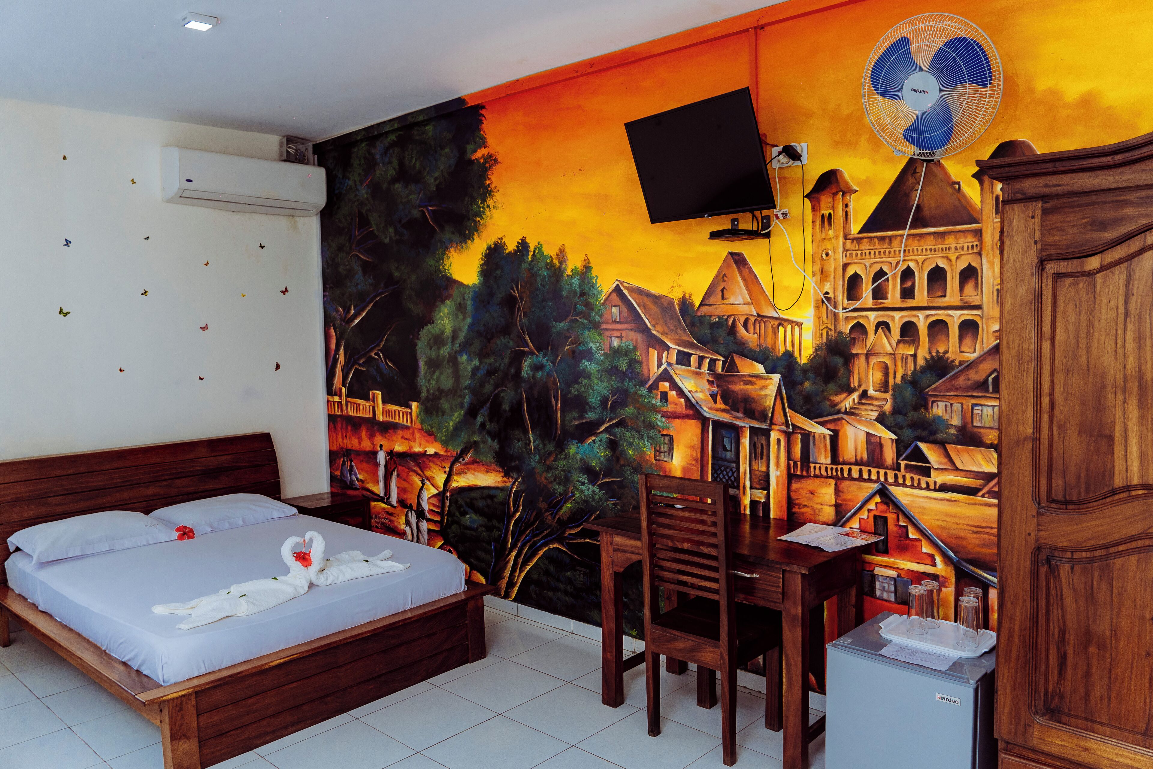 Une chambre d’hôtel ornée d’une fresque représentant un paysage de village, avec un lit recouvert de draps blancs, un bureau en bois et un petit réfrigérateur.