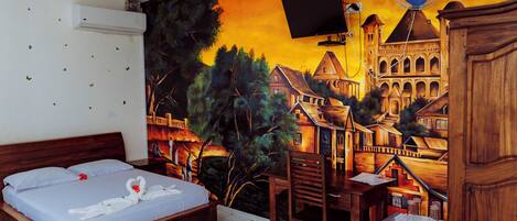 Une chambre d’hôtel ornée d’une fresque représentant un paysage de village, avec un lit recouvert de draps blancs, un bureau en bois et un petit réfrigérateur.