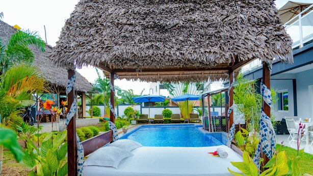 Una cama junto a la piscina con techo de paja, rodeada de abundante vegetación y una piscina.