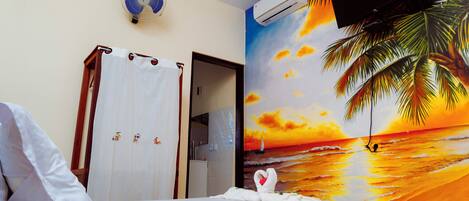 Ein Hotelzimmer mit einem Bett, einem Ventilator, einer Klimaanlage, einem Kleiderschrank und einem Wandbild mit einer Strandansicht.