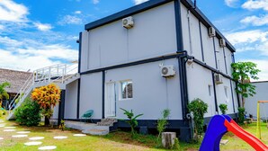 Un edificio de dos pisos con fachada blanca y detalles en negro, que presenta una escalera hacia la entrada, un ventanal pequeño y una unidad de aire acondicionado.