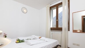 Une chambre d’hôtel propre et minimaliste, avec un lit, une table de chevet dotée d’une lampe, une fenêtre avec des rideaux et une vue sur les montagnes.