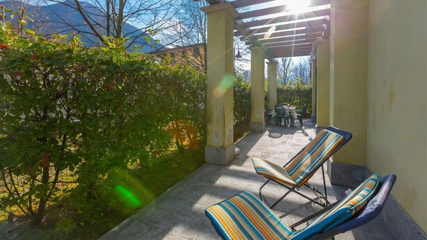 A covered patio with two striped lounge chairs, a pergola, and a view of mountains. - Porto Letizia Roma 1, Porlezza, Italy (Porlezza)