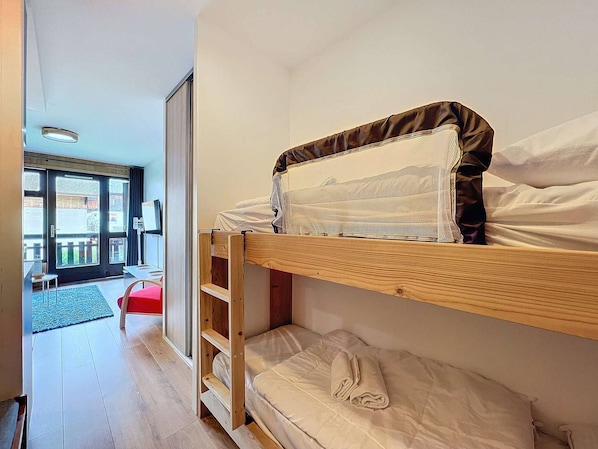 A bunk bed with a wooden frame and a mattress with a white cover. There is a small wooden shelf under the bed and a red chair in the room.