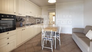 A modern kitchen with white cabinets, a built-in oven, and a dining area with a wooden table and white chairs. - L325 Appartamento - L325 PORTO MANNU (Palau)