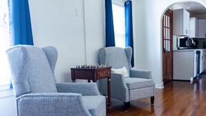 Een woonkamer met twee grijze fauteuils, een klein houten bijzettafeltje met een schaakspel, en een keuken op de achtergrond.