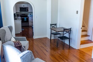 A living room with a sofa, a dining table with chairs, a kitchen in the background, and a staircase.