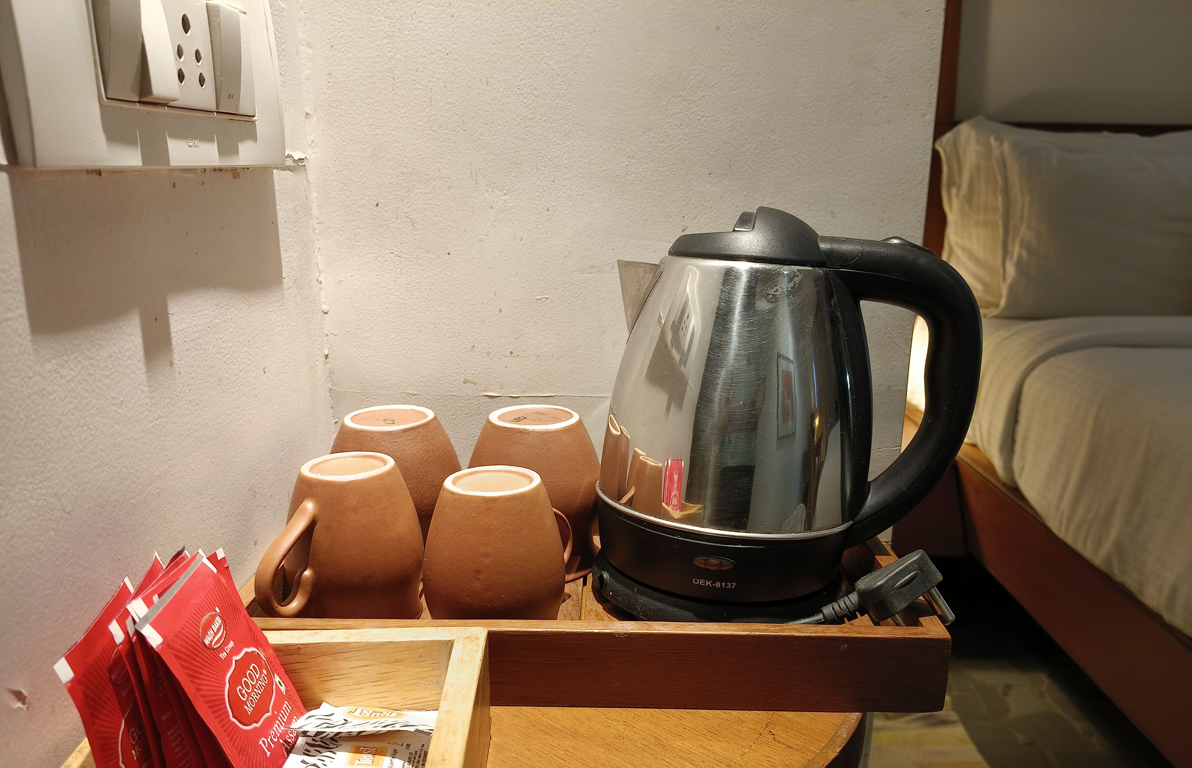 A kettle, several cups, and a tray with tea bags on a wooden table in a room with a bed and a wall socket.