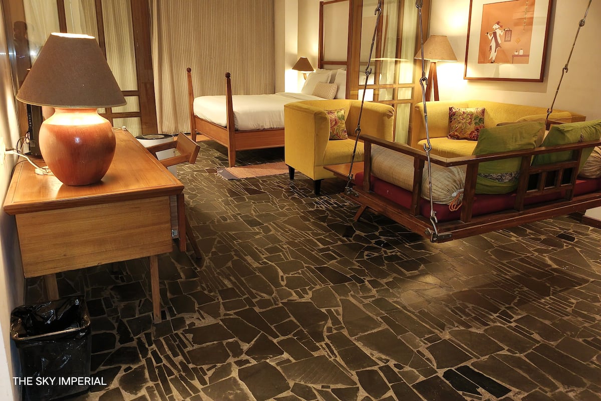 A hotel room with a bed, a sofa, a wooden table with a lamp, a hanging chair, and a stone-patterned floor.