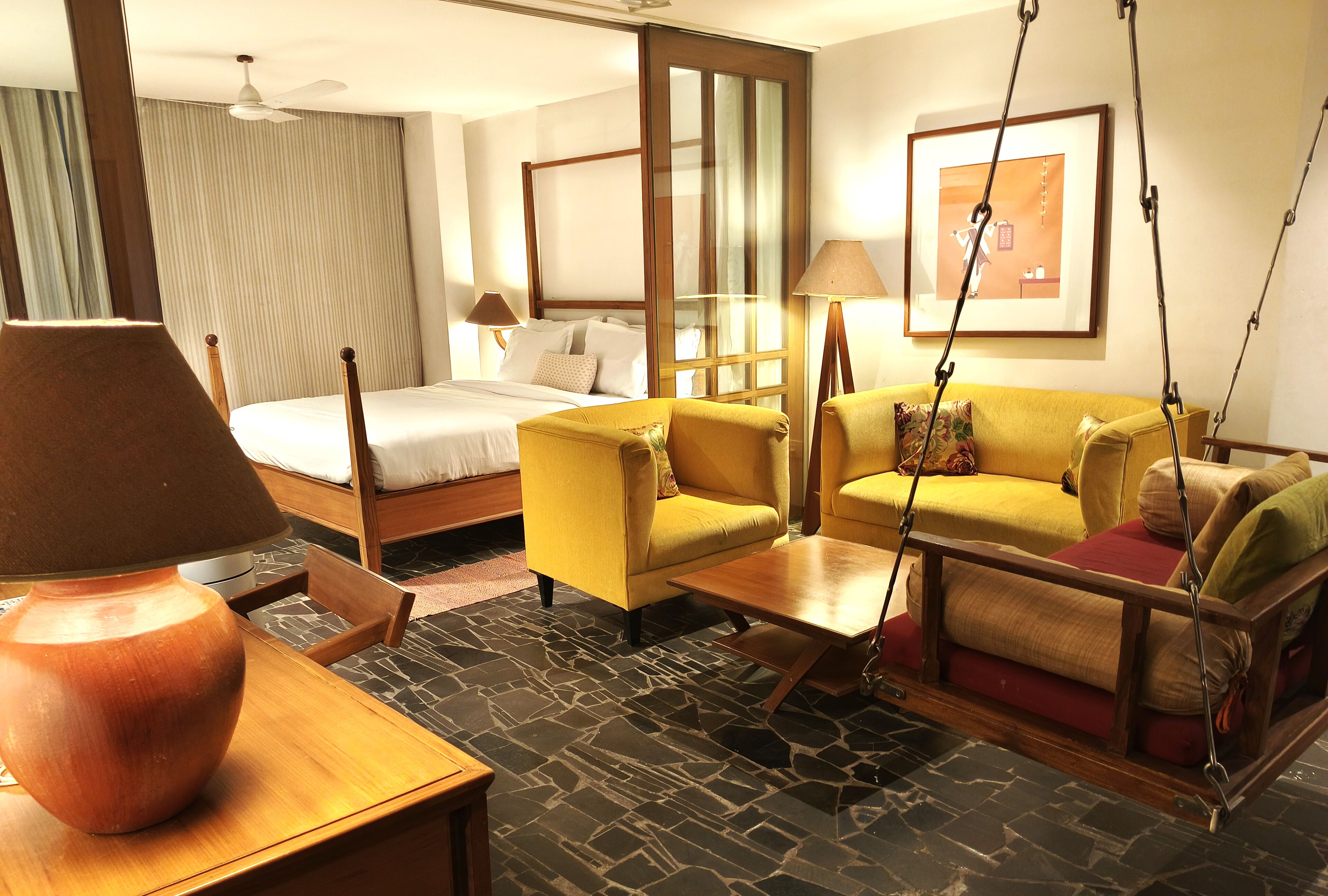 Habitación de hotel con cama, sofá amarillo, mesa de centro de madera, sillón colgante y un espejo grande.