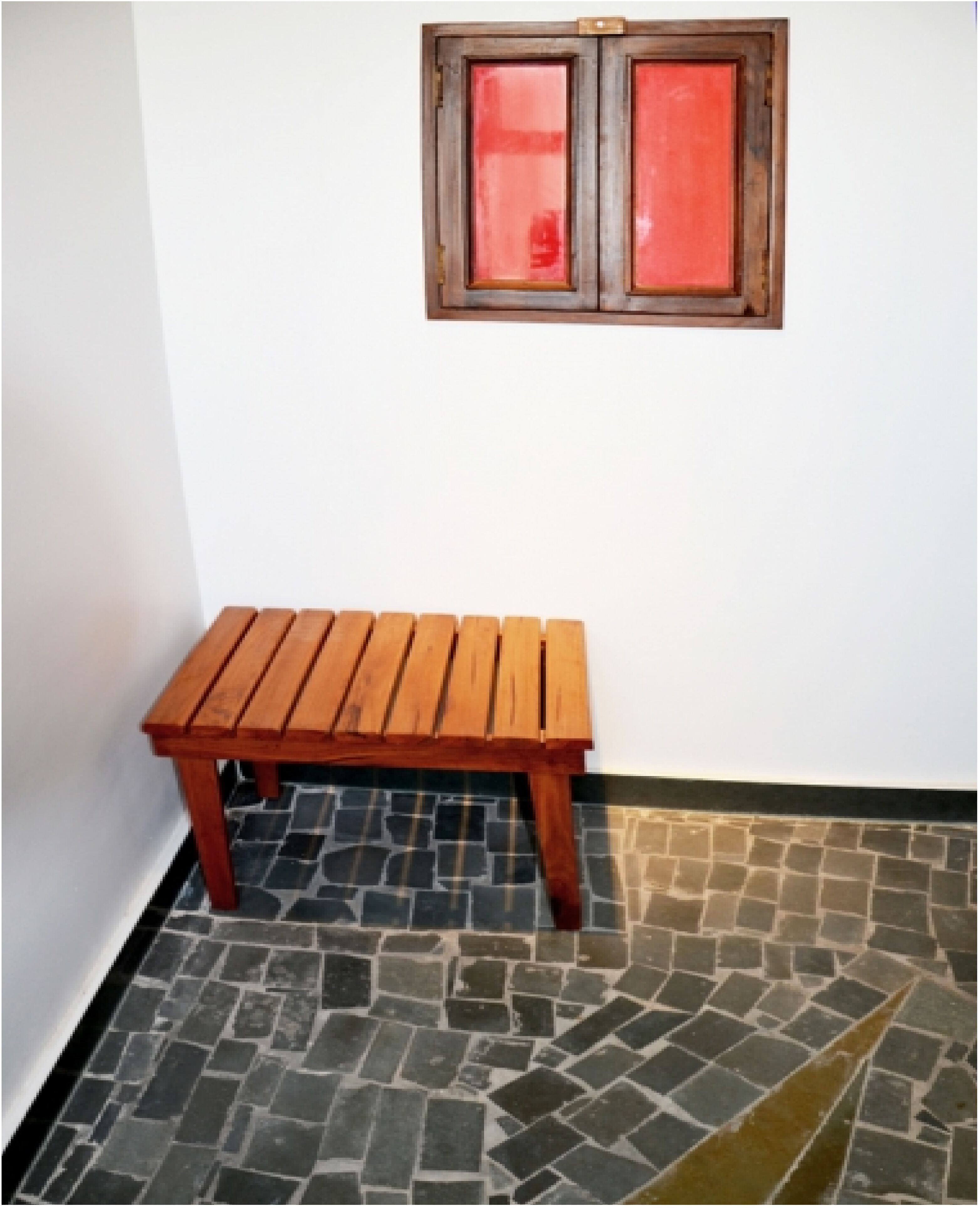 Eine hölzerne Bank vor einer weißen Wand mit einem Fenster darüber.